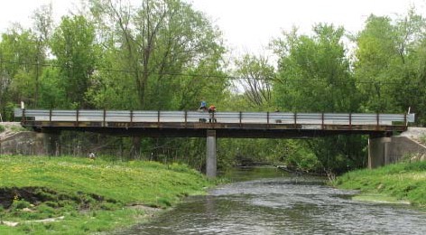An image of pedestrians on a rural bridge, lined with trees on either side, looking at the stream below.